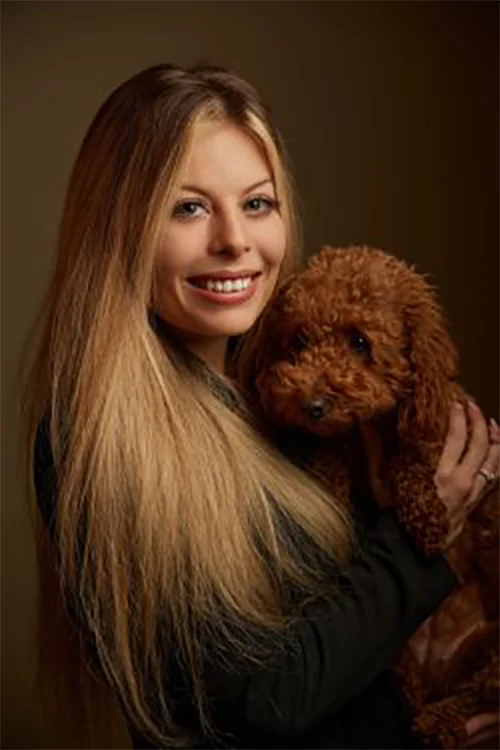 A person with long blond hair smiles while holding a small brown curly-haired dog.