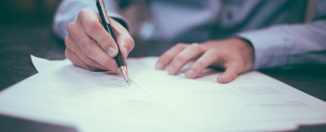 A person is writing on multiple sheets of paper with a pen, wearing a long-sleeve shirt, seated at a desk.