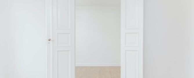 An open white double door reveals an empty room with white walls and light wooden flooring.