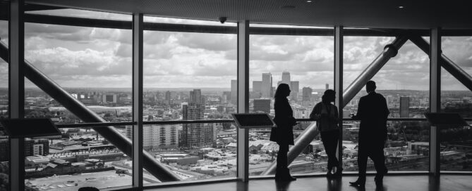 Three people stand near large windows overlooking a city skyline from a high-rise building. The room has a modern design with visible structural beams.