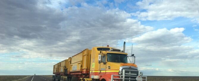 A large yellow road train truck with multiple trailers is driving down a long, empty highway under a partly cloudy sky.