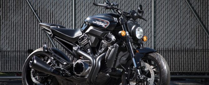 A black motorcycle with silver accents is parked on a pavement in front of a tall, industrial metal fence.