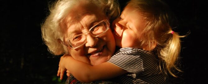 An elderly woman with glasses smiles as a young girl with blonde hair hugs and kisses her on the cheek in a warmly lit setting.