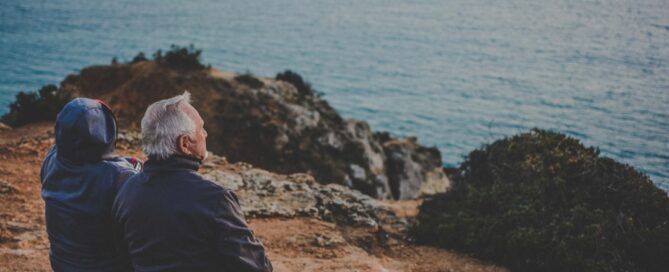 Two elderly individuals sit on a rocky cliff, overlooking a vast body of water.