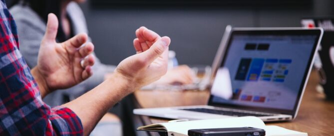 Two individuals sit at a conference table, one gesturing with hands while the other uses a laptop. The table has a notebook, phone, and a laptop displaying various charts.