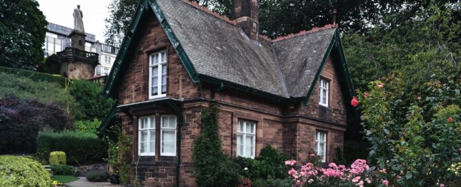 A quaint brick cottage with a steeply pitched roof is surrounded by a lush garden with various flowers and shrubs, set against a backdrop of tall trees and a statue in the distance.