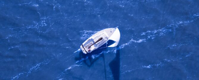 Aerial view of a sailboat sailing on a blue sea with visible waves and a shadow of the mast on the water.