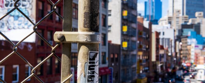 View of a city street from behind a chain-link fence, with various buildings and signs in the background under a clear blue sky.