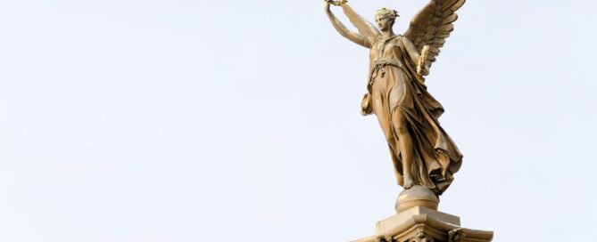 A stone statue of a winged angel holding a wreath, standing on top of an ornate column against a clear sky.