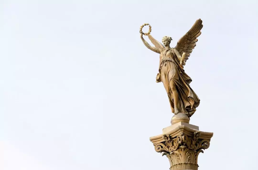 A stone statue of a winged angel holding a wreath, standing on top of an ornate column against a clear sky.