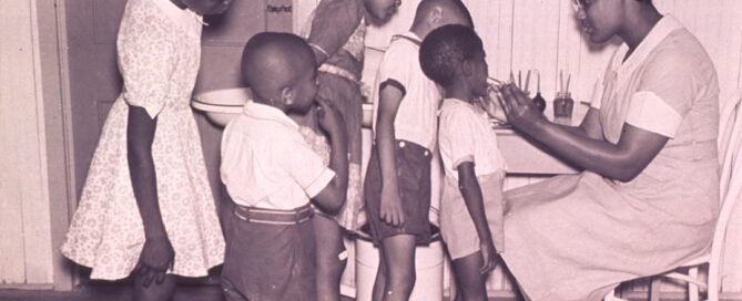 A woman sits at a table checking the health of four children standing in a line. Another child stands nearby in a dress. The room has wooden walls and flooring.