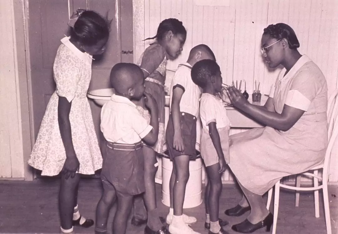 A woman sits at a table checking the health of four children standing in a line. Another child stands nearby in a dress. The room has wooden walls and flooring.