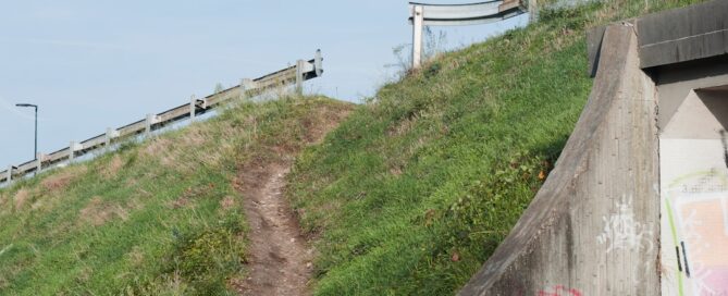 A dirt path runs up a grassy slope alongside a guardrail near a concrete structure with graffiti.