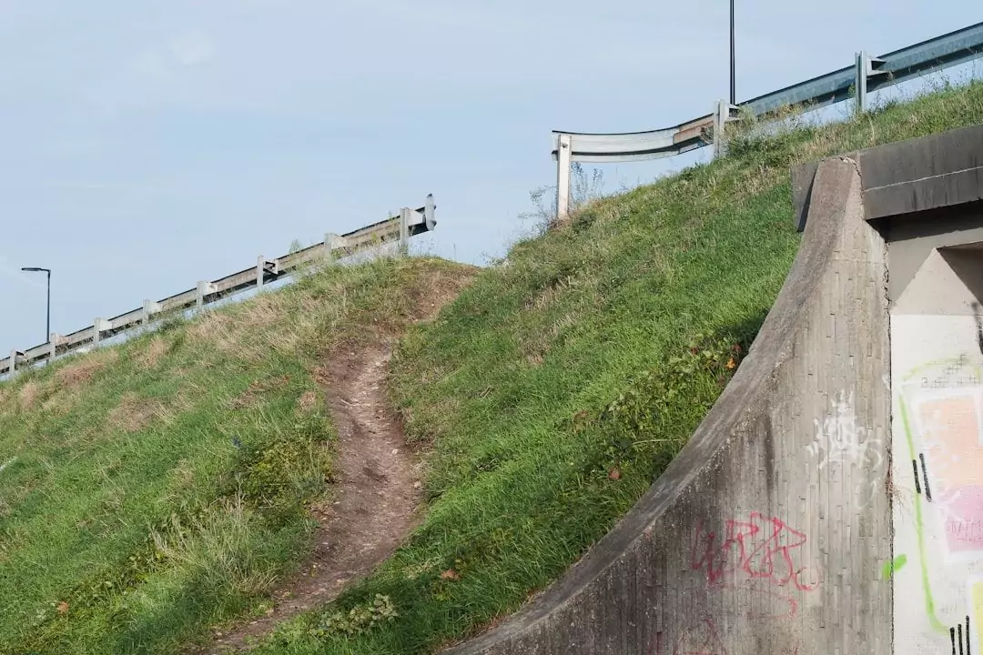 A dirt path runs up a grassy slope alongside a guardrail near a concrete structure with graffiti.