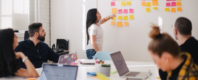 A woman stands at a whiteboard covered in sticky notes, presenting to four seated colleagues during a meeting in a bright office space.