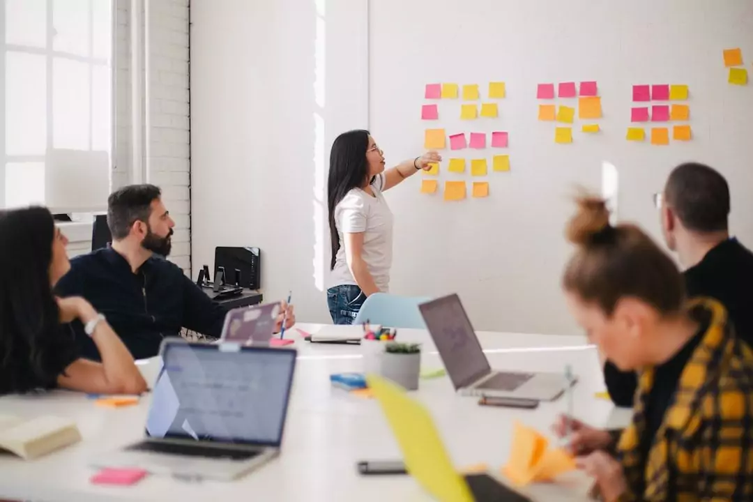 A woman stands at a whiteboard covered in sticky notes, presenting to four seated colleagues during a meeting in a bright office space.