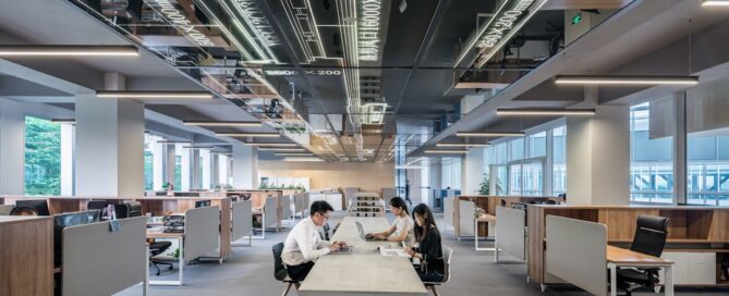 A modern open office space with people working at desks and sitting at a central table under mirrored and illuminated ceiling panels.