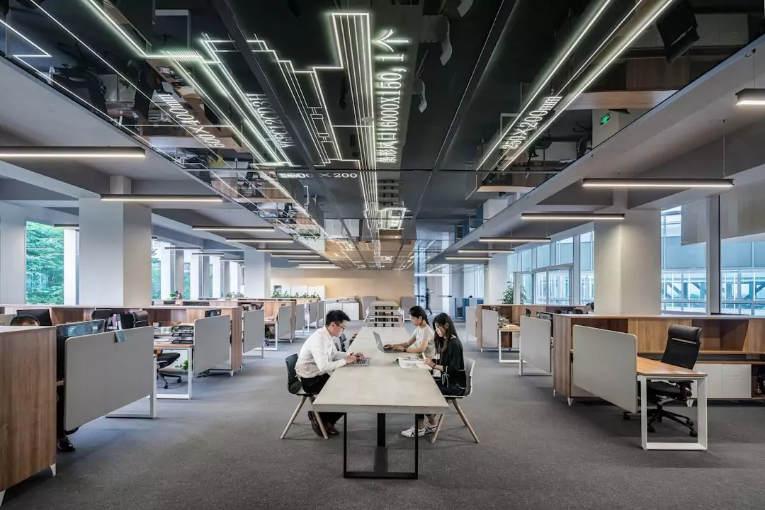 A modern open office space with people working at desks and sitting at a central table under mirrored and illuminated ceiling panels.