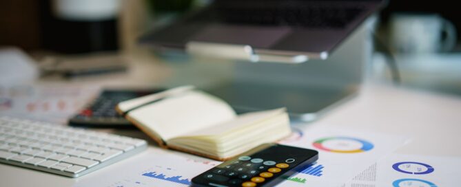 A workspace with a keyboard, a notebook, a calculator, and financial charts on paper. A laptop is on a stand in the background.