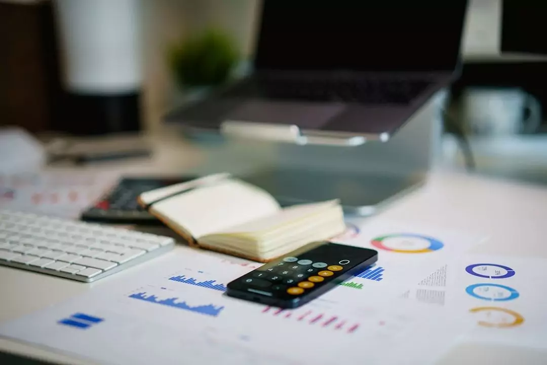 A workspace with a keyboard, a notebook, a calculator, and financial charts on paper. A laptop is on a stand in the background.