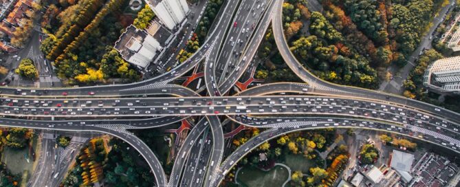 Aerial view of a busy multi-layered highway interchange surrounded by densely packed buildings and lush greenery. Traffic flows smoothly across several lanes and bridges.