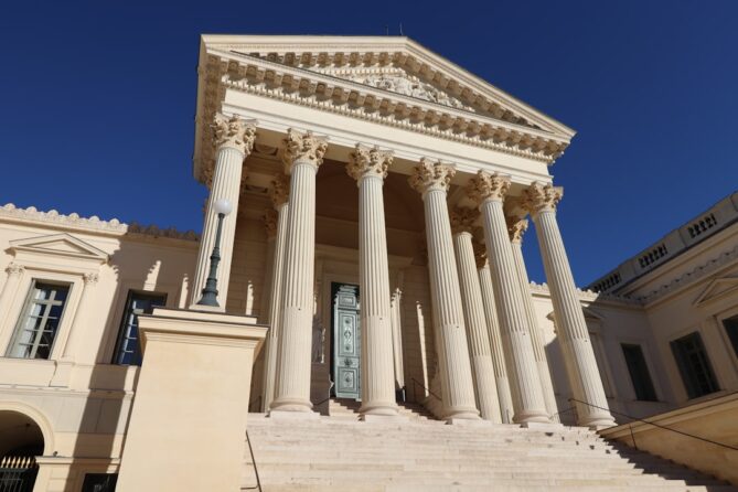 Grand neoclassical building with tall columns and a triangular pediment, featuring a central entrance door, set against a clear blue sky.