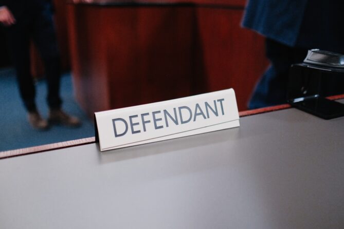 A close-up of a sign labeled "Defendant" on a table in a courtroom setting.