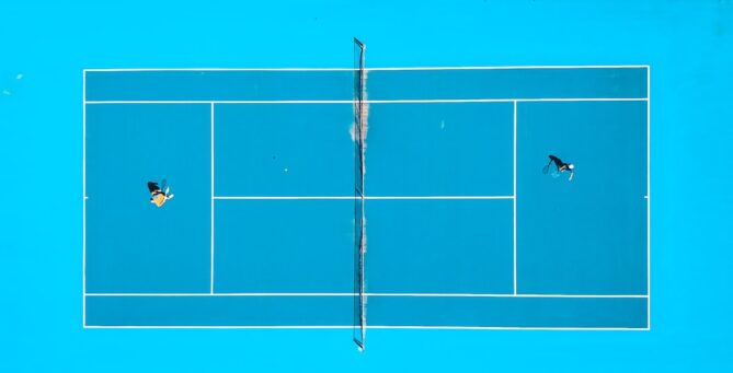 Aerial view of a tennis court with two players competing. The court has a bright blue surface and white lines. A net divides the court in the middle. One player is at each side of the net.