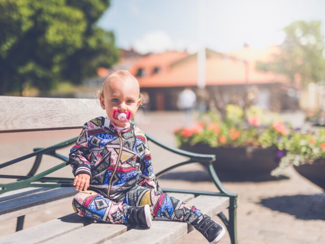 A toddler with a pacifier sits on a bench outside, dressed in a colorful, patterned outfit. The background features blurred buildings and plants in bright daylight.