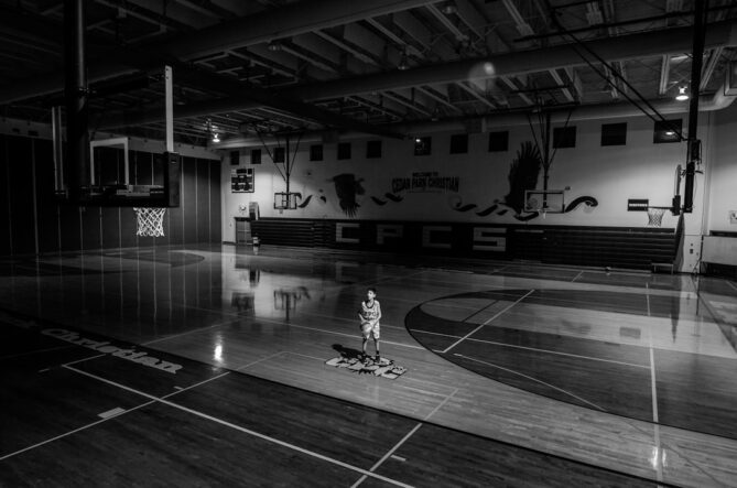 Black and white image of an empty basketball court with a single player holding a ball at center court, illuminated by a spotlight. The gym has banners and bleachers in the background.