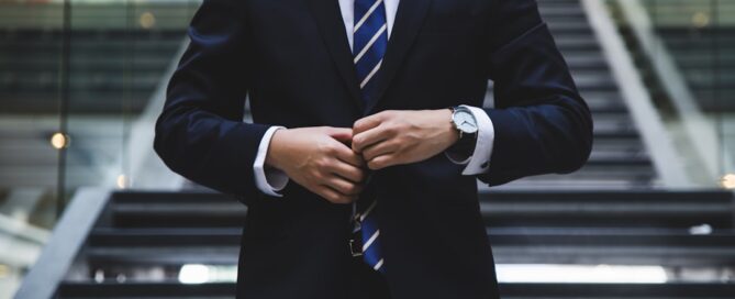 A person in a suit and tie stands in front of modern stairs, buttoning their jacket.