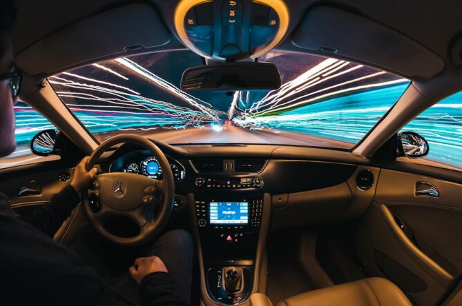 View from inside a car driving at night with blurred light trails outside the windshield. The dashboard and steering wheel are visible, and a person is seated behind the wheel.