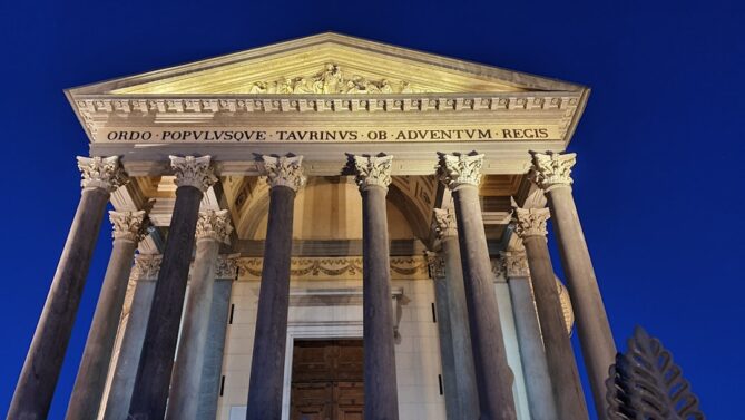 Front view of a historical building with tall columns and a triangular pediment at night, featuring an inscription in Latin across the top.