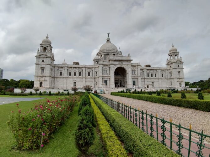 A grand white stone building with intricate designs, featuring large domes and towers, set against a cloudy sky. Manicured gardens and pathways lead up to the entrance.