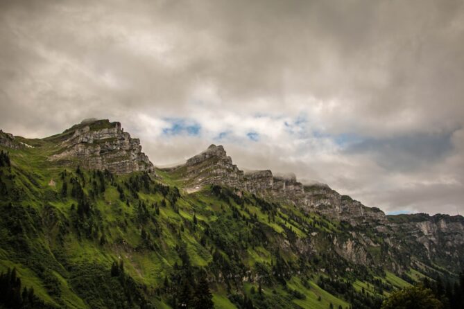 A rugged mountain range with steep, rocky peaks and lush green slopes under a cloudy sky.