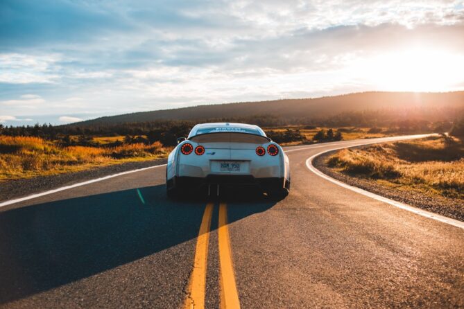 A white sports car is driving down a winding road with a scenic landscape and a bright sun in the background.