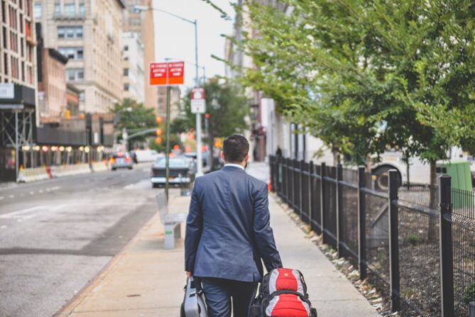 A man in a suit walks down a city sidewalk carrying a briefcase and a suitcase, with trees and buildings lining the street.