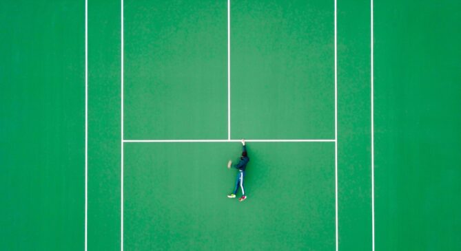 A person lies on their back in the middle of a green tennis court, with one arm stretched out and legs slightly bent.