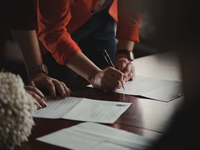 Person in a red jacket is signing a document on a wooden table with scattered papers around, while another person observes.