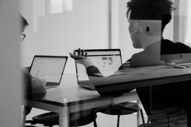 Two people are working at laptops in an office setting, viewed through a glass partition. One person gestures towards their screen in discussion.