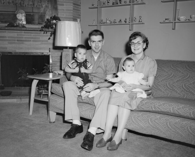 A man and woman sit on a couch, holding two young children. The room features a fireplace, a table lamp, and decorative figurines on shelves.