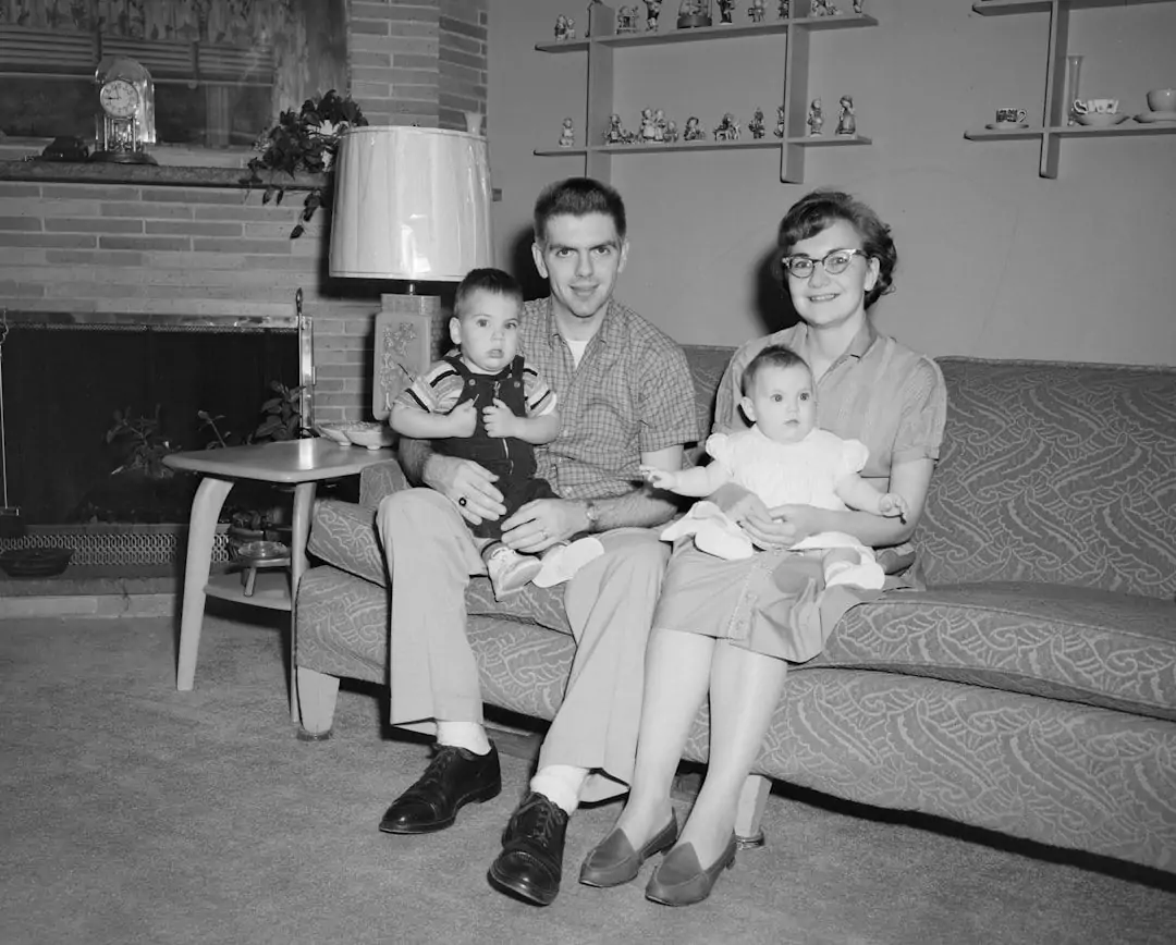 A man and woman sit on a couch, holding two young children. The room features a fireplace, a table lamp, and decorative figurines on shelves.