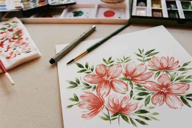 A painting of pink and red flowers with green leaves on a white paper, surrounded by paintbrushes, a pen, and watercolor palettes.