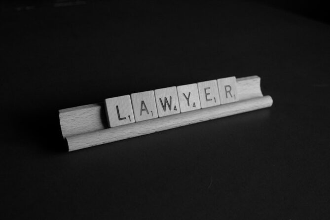 Black and white image of Scrabble tiles spelling out the word "LAWYER" on a wooden tile rack against a dark background.