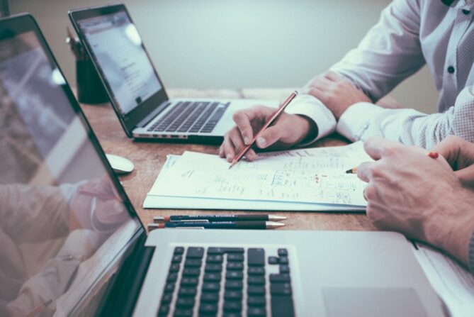 Two people work on documents at a table with laptops, pencils, and papers in a collaborative office setting.