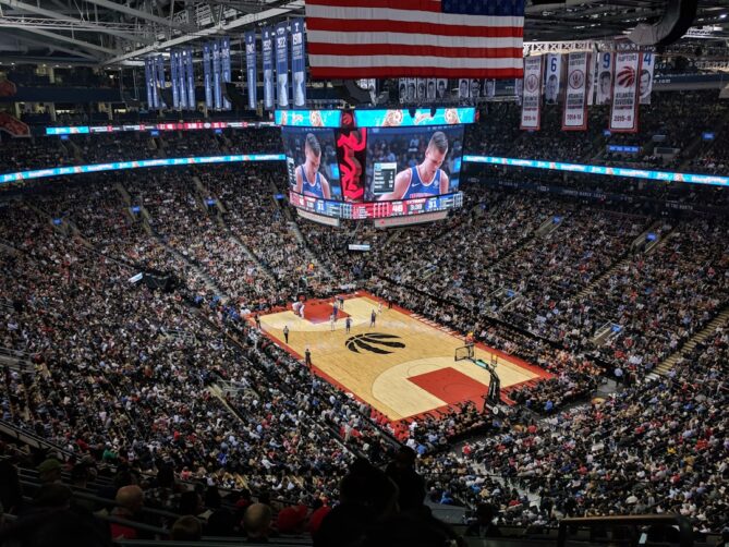 A packed arena during a basketball game, featuring a large scoreboard displaying close-up footage of the players on the court. The court has a distinctive black logo at its center.