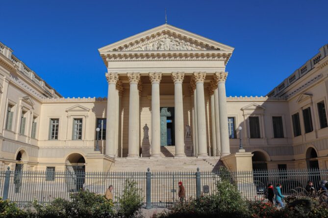 Front view of a neoclassical building with large columns and a classical pediment. People are walking in front of it, and a clear blue sky forms the background.