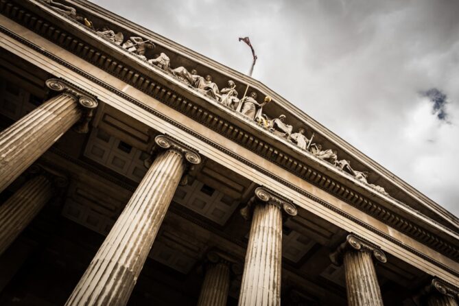 A low-angle view of a neoclassical building with tall columns and sculpted figures along the top, under a cloudy sky.