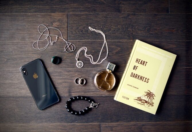 A black iPhone, perfume bottle, rings, necklace, black bracelet, earphones, and the book "Heart of Darkness" by Joseph Conrad on a dark wooden surface.