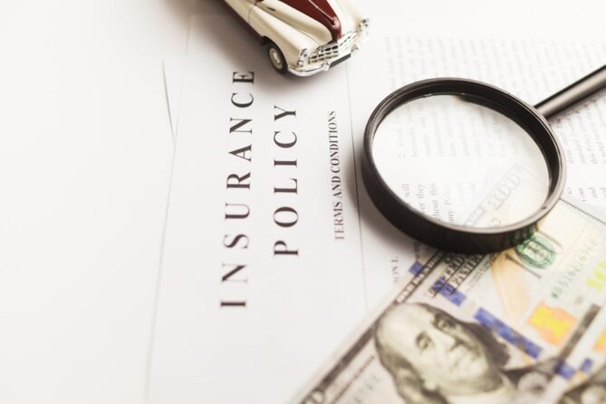 A magnifying glass and toy car rest on top of an insurance policy document and a hundred-dollar bill.